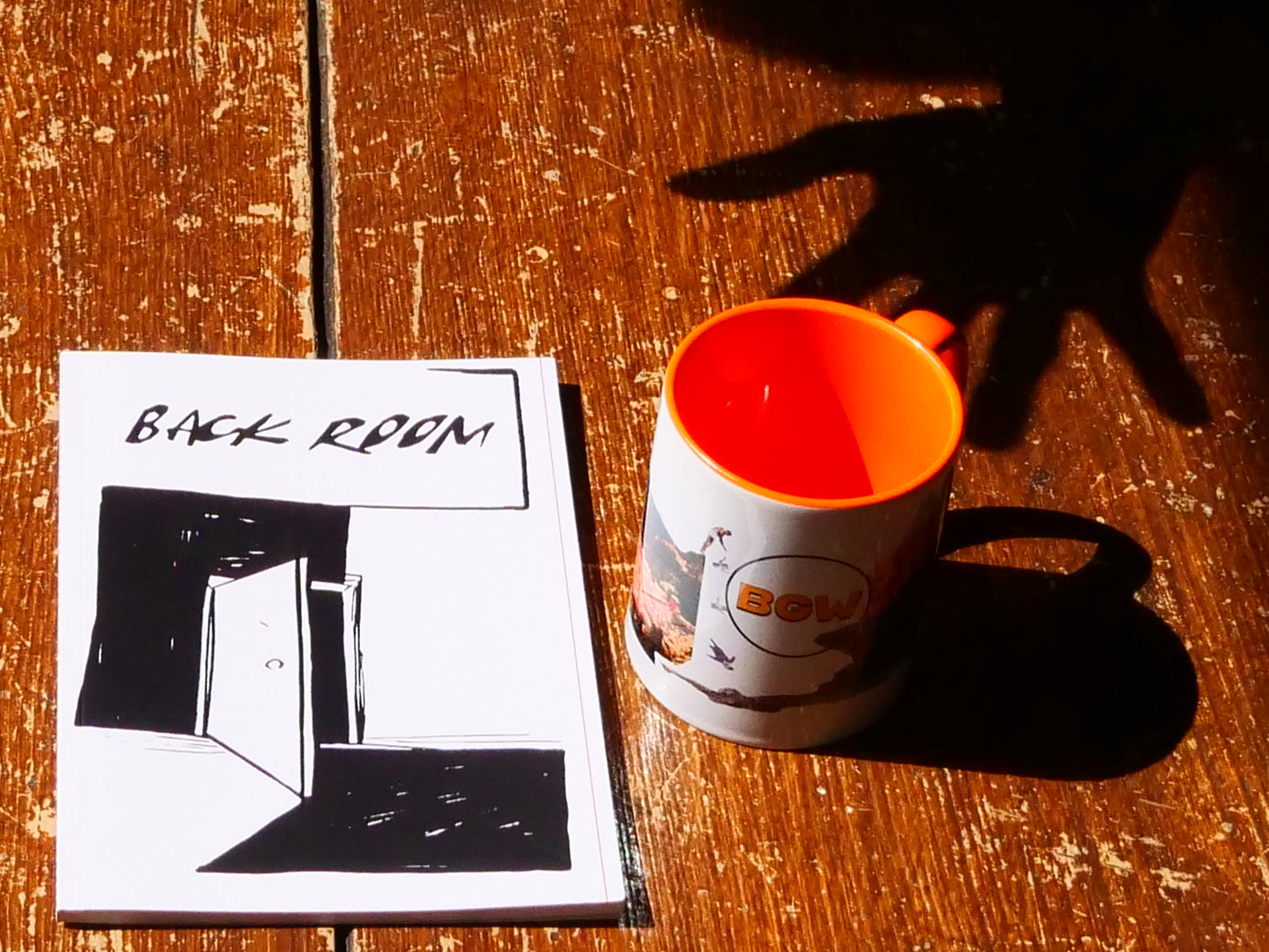 A white ceramic mug with a bright orange interior and handle sits on a rustic wooden table next to a printed booklet titled 'BACK ROOM' with black line art. Strong sunlight casts a hand-shaped shadow across the table.