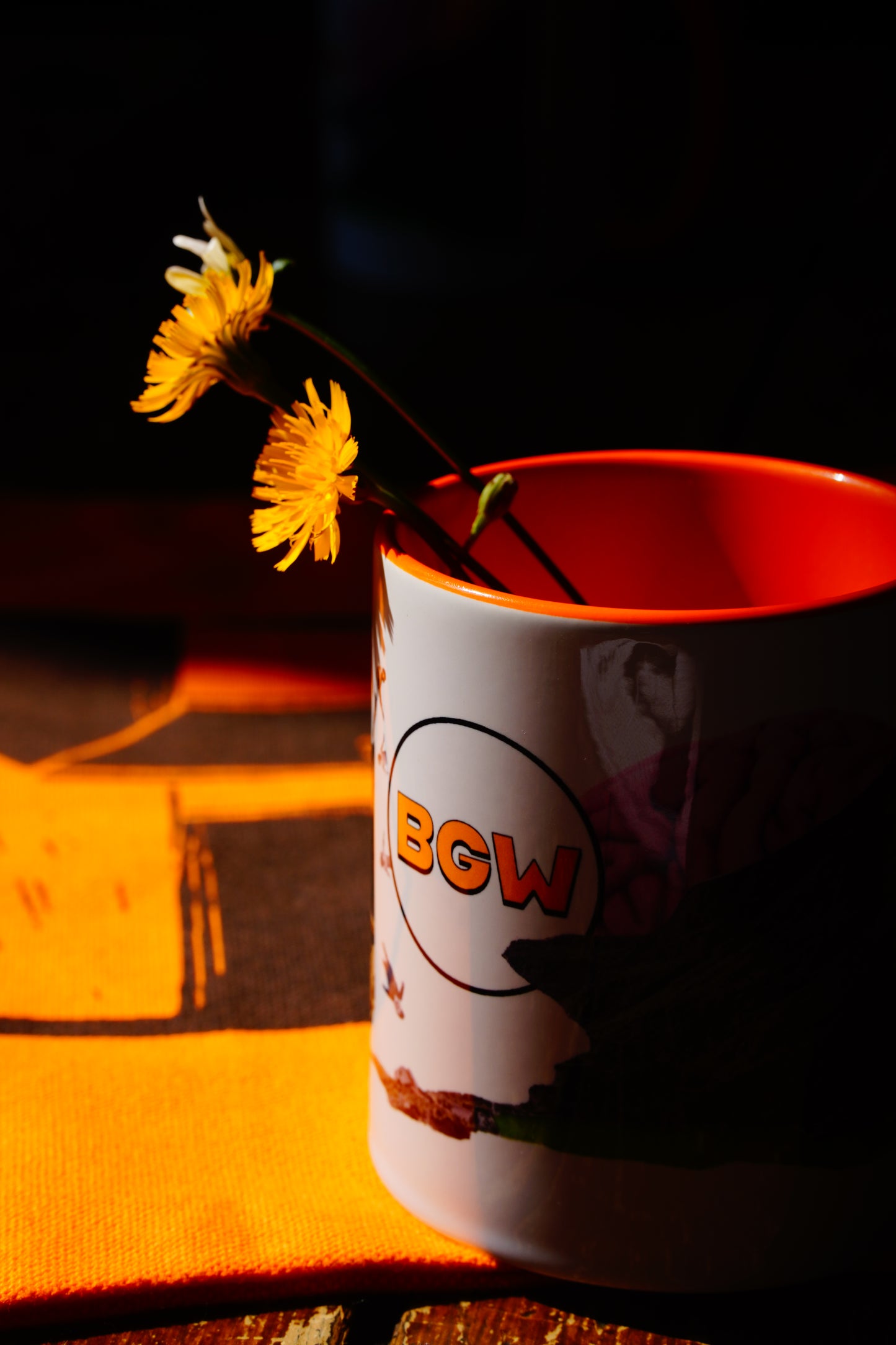 A white ceramic mug with a bright orange interior, featuring a graphic print and the text 'BGW' in orange, holding two yellow flowers against a dark, moody background with dramatic warm lighting.