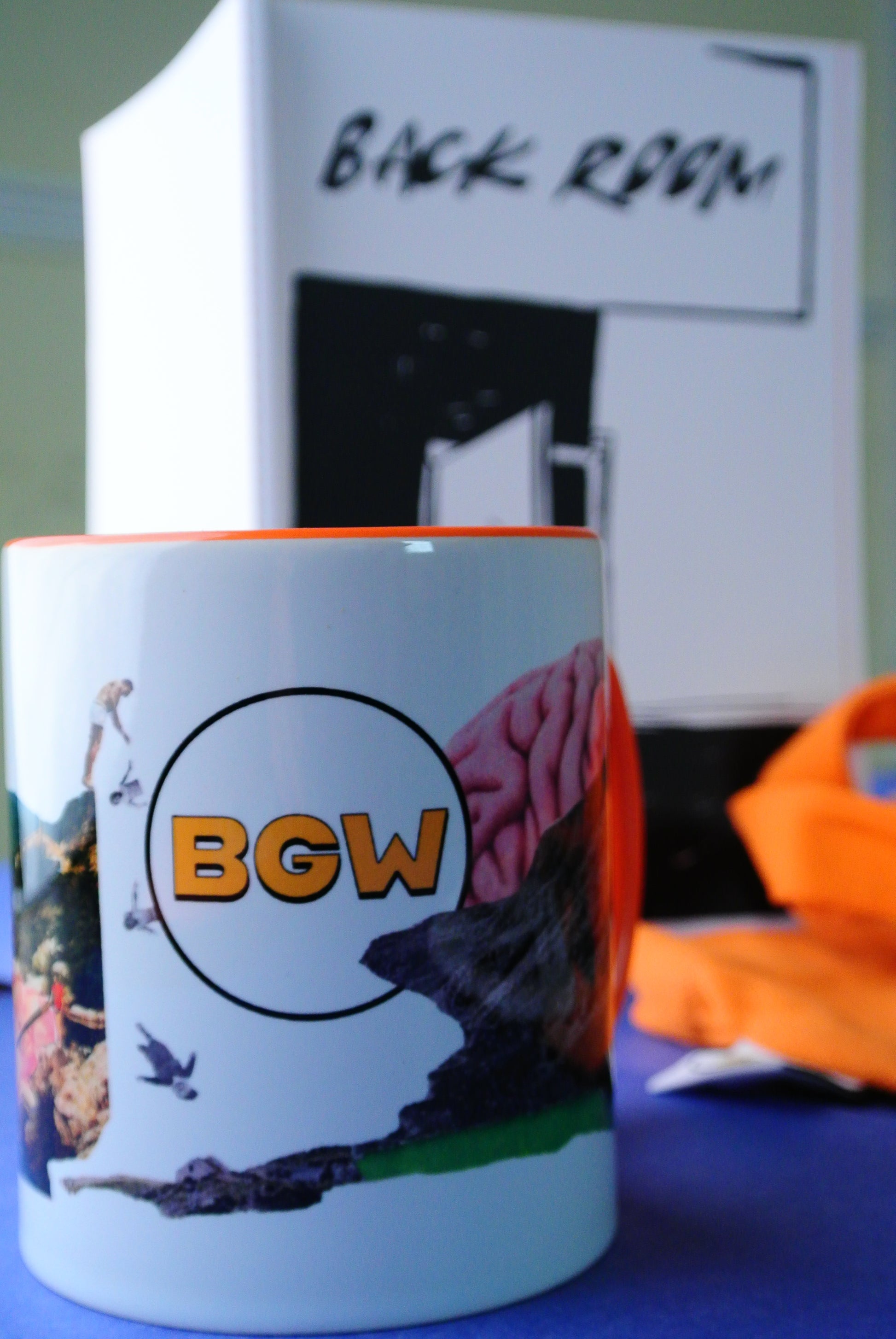 A white ceramic mug with a colorful graphic print and the letters 'BGW' in orange inside a black circle, shown next to a white notepad with a cover text of 'BACK ROOM' just out of focus behind.