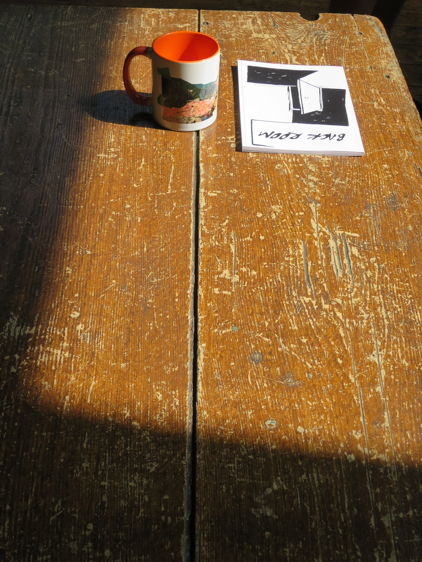 A small ceramic mug with a bright orange interior and handle is mounted on a weathered wooden surface next to a small printed notebook.