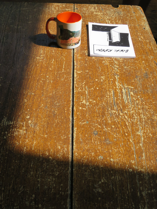 A small ceramic mug with a bright orange interior and handle is mounted on a weathered wooden surface next to a small printed notebook.