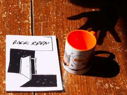 A white ceramic mug with a bright orange interior and handle sits on a rustic wooden table next to a printed booklet titled 'BACK ROOM' with black line art. Strong sunlight casts a hand-shaped shadow across the table.