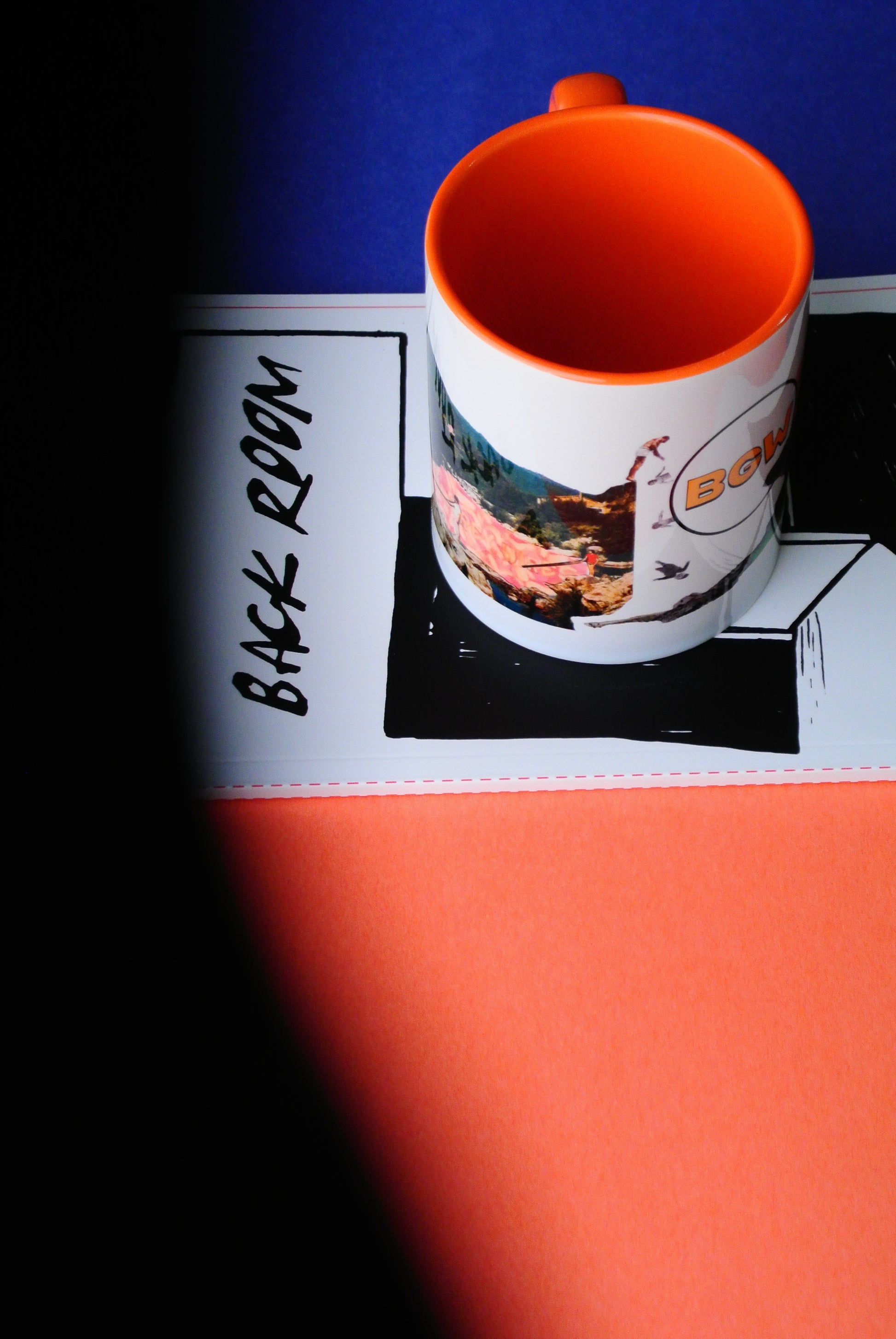 A white ceramic mug with a bright orange interior and handle, featuring a graphic print that was collaged, resting on a notepad with the text 'BACK ROOM', sits against a split blue and orange background.