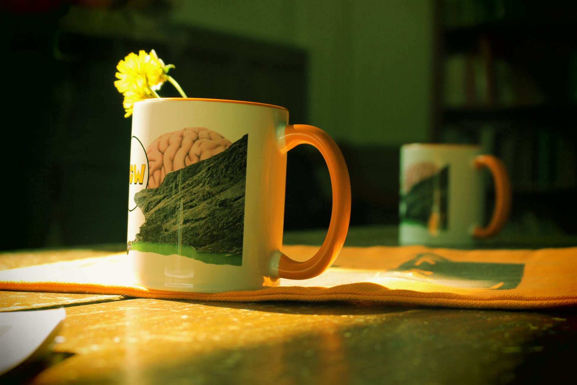A white ceramic coffee mug with an orange handle and a printed scenic graphic, set on a wooden table with a small yellow flower in a warm, low-light environment.