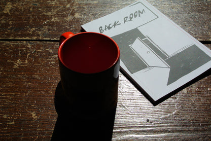 A mug with an orange interior and handle, placed on a rustic wooden table next to a printed notebook labeled 'BACK ROOM' with a black-and-white illustration.