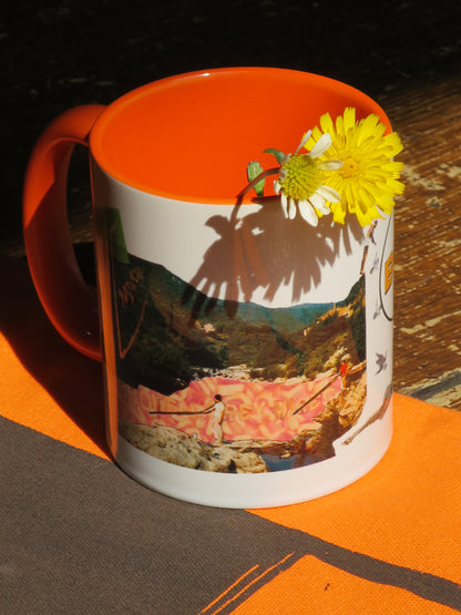 A white ceramic mug with a bright orange handle and interior, featuring a printed scenic photograph on the side. Two small flowers rest inside the orange rim, casting a shadow. The mug is photographed on an orange and dark grey printed tote bag.